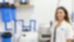 Smiling woman in a SoCal Water Solutions shirt stands by water filtration equipment with blue filters and tubes in a clean, white room.
